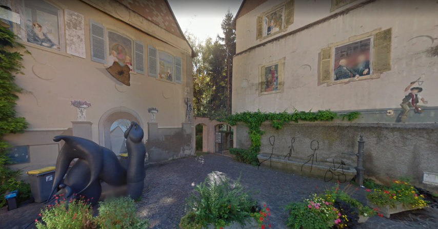 Une capture de street view à l'emplacement de la fontaine, la rue est pavée, il y a des plantes vertes et du lière, une statue de silhouettes rondes humaines, une petite famille est représentée. Sur les murs, des trompes l'oeil sont peints, avec des fenêtres ouvertes sur des personnages et leur quotidien. J'ai dessiné grossièrement trois stick-human installé sur la fontaine, et un vélo.